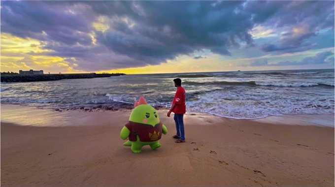 A boy in a red jacket stands on a beach at sunset facing a large inflatable green dragon mascot character, with the ocean and cloudy sky in the background.