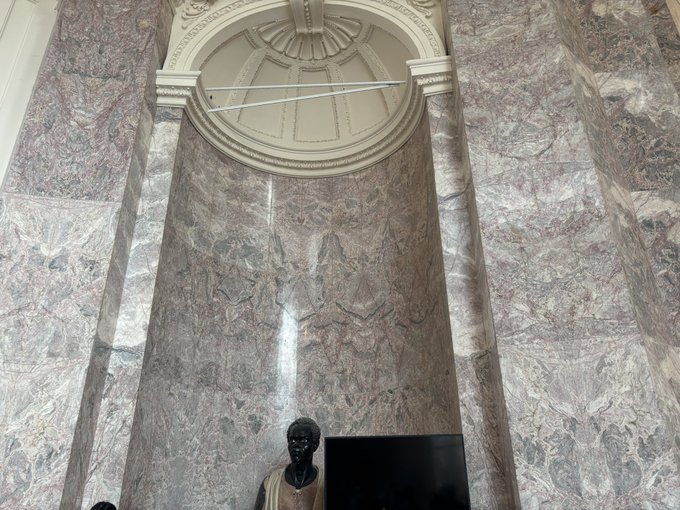 An interior view of an ornate building featuring tall columns made of purple and pink veined marble, with decorative white molding and a domed ceiling visible at the top. A dark bust statue and the top of a monitor or screen are visible at the bottom of the frame.