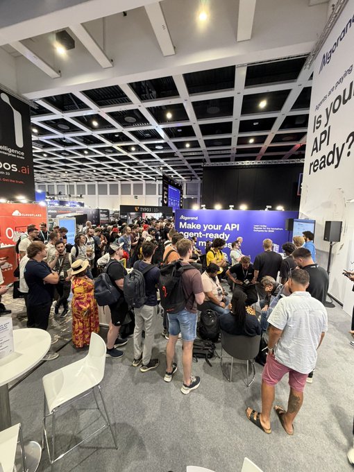A crowded indoor event or convention with many attendees standing and talking; a large blue sign in the background reads 'Make your API agent-ready' and a banner asks 'Is your API ready?'.
