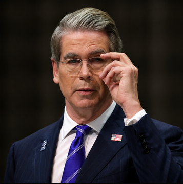 A photograph of a man in a navy suit, blue striped tie, and American flag pin, adjusting his glasses.