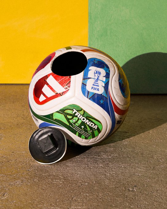 An Adidas FIFA Trionda soccer ball resting on the ground against a yellow and green background, with a black cap in front of it.