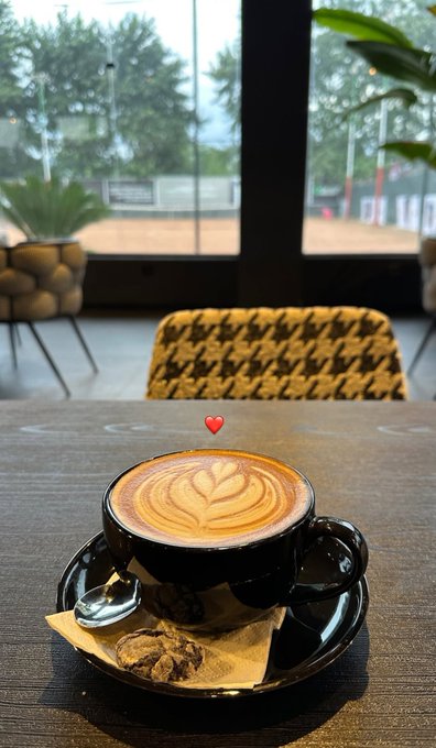 A heart-shaped latte art cappuccino sits on a wooden café table beside a cookie, with a tennis court visible through the window — a cozy weekend morning vibe paired with the post's shoutout to the @0xAlphaGEMs community by @CrypSaf.
