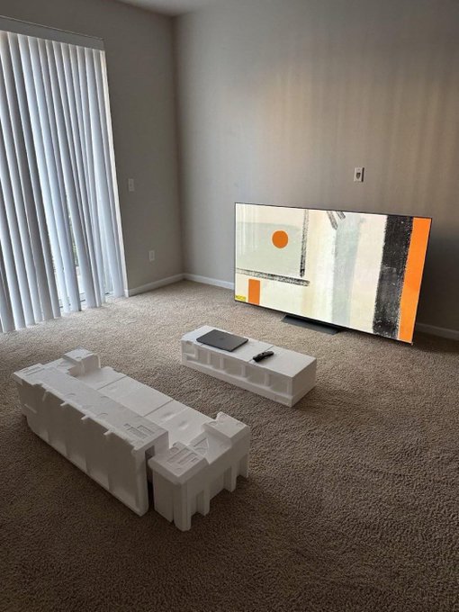 Living room with beige carpet, vertical blinds, a large TV displaying abstract artwork, and two white sculptural block-style coffee tables.