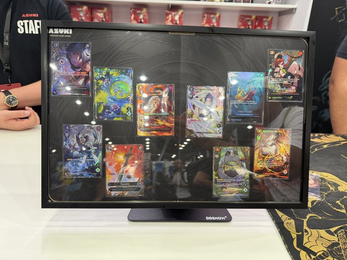 A staff member's lanyard partially visible at a convention displays "AZUKI STAFF" text, while a DDSMOM display case showcases a collection of holographic Azuki trading cards featuring various anime-style characters and artwork.