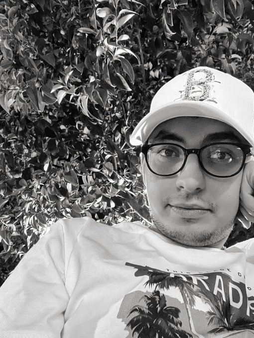 A black-and-white selfie of a young man wearing glasses and a cap, posed in front of foliage.