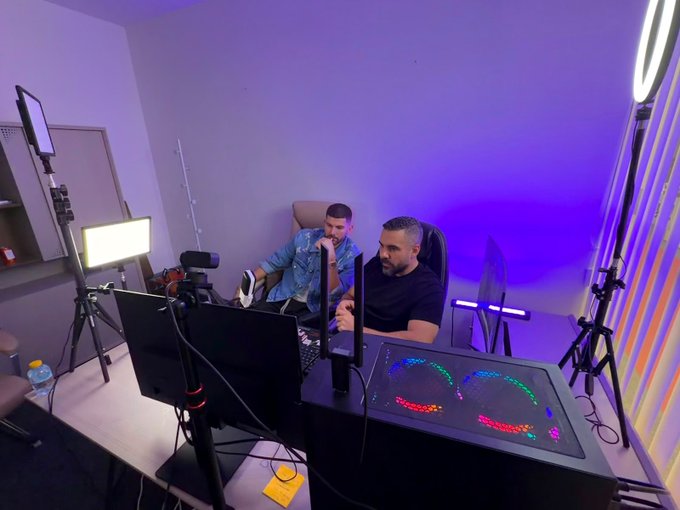 Two men are seated at a studio desk equipped with multiple monitors, cameras, and colorful RGB lighting, appearing to record a podcast or livestream session that aligns with the post's promise of sharing crypto alpha insights.