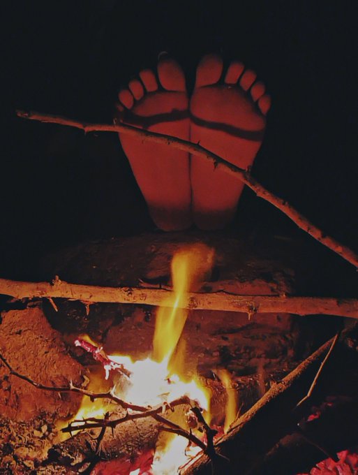 A pair of feet illuminated by the glow of a campfire with sticks burning in the foreground.