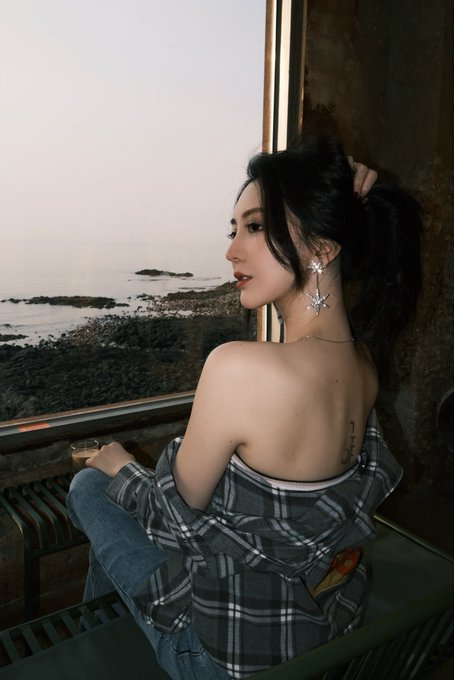 A young woman with dark hair in a plaid shirt sits by a window looking out at a coastal landscape, revealing a back tattoo and star-shaped earrings.