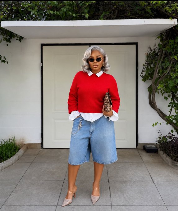 A woman with silver hair, sunglasses, a red sweater, and denim Bermuda shorts poses in front of a white door, holding a leopard-print bag.