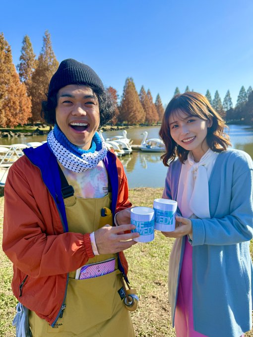 Two people smiling outdoors by a lake, holding ONE MEDY branded cups, dressed in colorful outdoor clothing with autumn trees in the background.