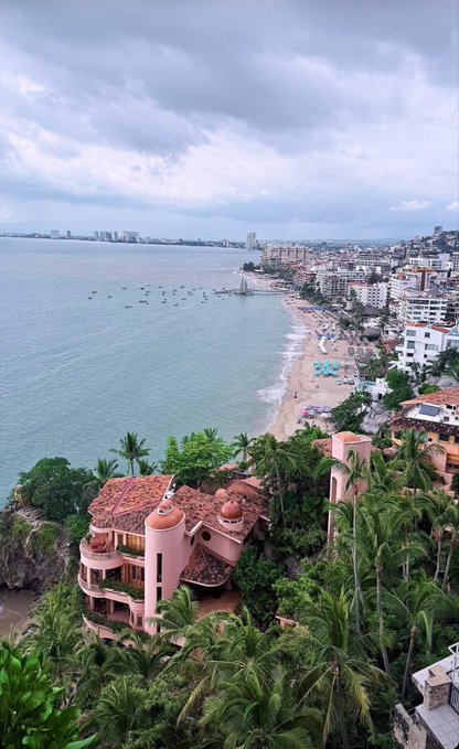 An aerial view of a coastal city with a pink, dome-roofed building nestled among palm trees in the foreground, overlooking a beach and the ocean under a cloudy sky.