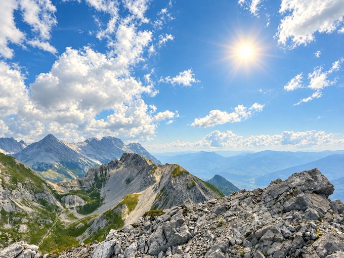A panoramic view from a rocky mountain peak shows jagged ridges, a valley, and distant mountain ranges under a bright sun in a partly cloudy blue sky.