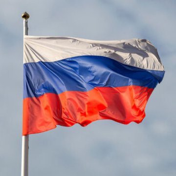 The Russian flag (white, blue, red horizontal stripes) waves on a flagpole against a cloudy sky.