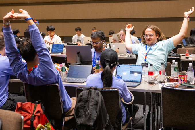 A group of young people wearing blue shirts work on laptops at a conference table, with several raising their arms in celebration.