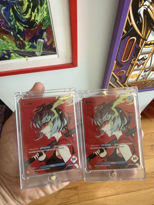 A hand holds two identical 'Shaq' trading cards encased in protective sleeves, featuring an anime-style character with red and teal hair, displayed in front of colorful framed artwork.
