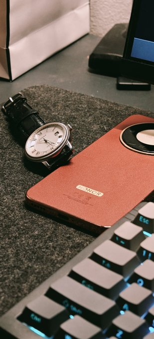 A desk setup featuring a leather-strapped wristwatch, an orange/salmon smartphone, and a backlit mechanical keyboard on a gray felt surface.