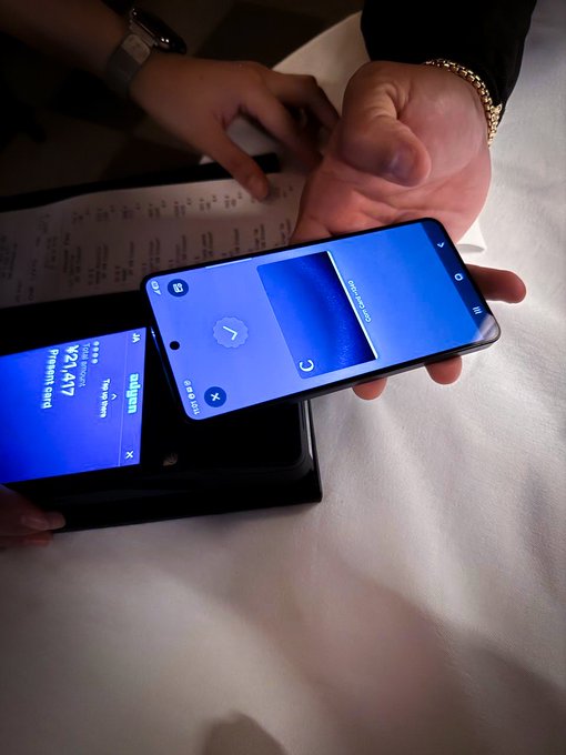 A person holding two smartphones, one displaying a camera app with 'Camera in use' and another showing the SoDEX app with 'TOTAL Holdings W2:1,417' and '77,157' SoPoints.