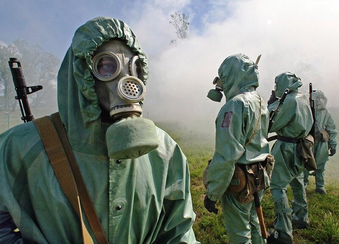 Three individuals wearing full-body protective hazmat suits and gas masks stand in a smoky field, appearing to be conducting training or emergency response operations. The image humorously contrasts the extreme protective gear with the caption's complaint about having to work, suggesting that hazardous conditions aren't enough to warrant a day off.