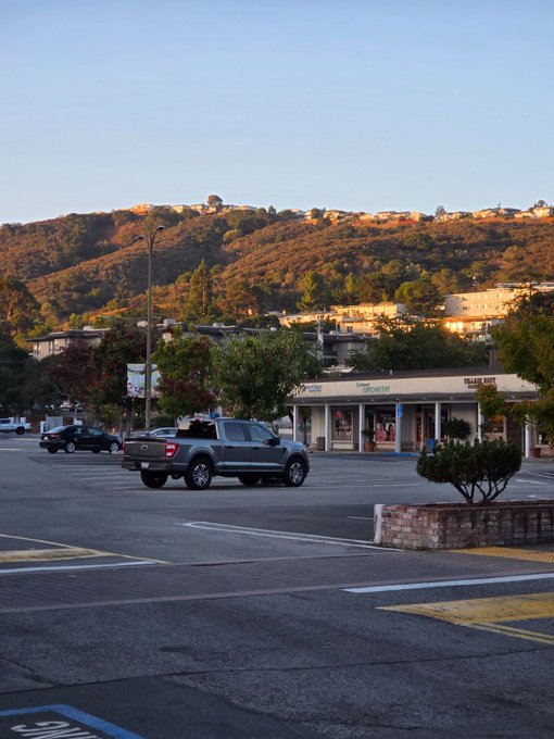 A gray pickup truck parked in a lot during the day, with hills and storefronts visible in the background.