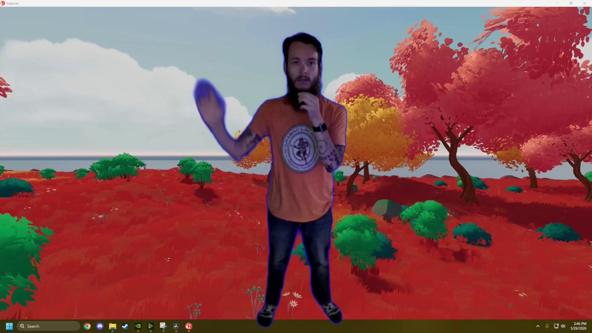 The image shows a man standing in front of a computer screen, wearing an orange shirt and jeans. The background features a cartoonish scene of trees and bushes rendered in red and green colors. The text "This is what is looks like to build in @decentraland these days Walkthrough with explanations for a new builder!" is displayed above the man's head, suggesting that he might be participating in a virtual walkthrough or tutorial related to building in the game Decentraland.