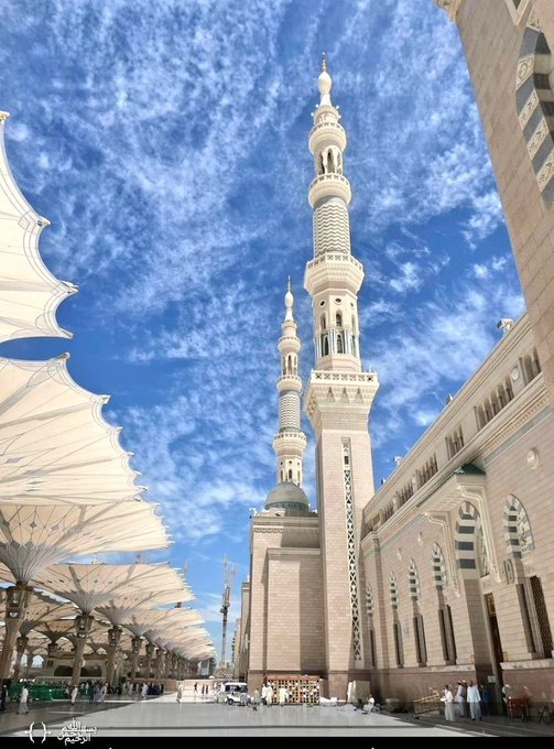 A tall mosque minaret reaching into a blue cloudy sky, with distinctive white umbrella-like shade structures in the courtyard below.