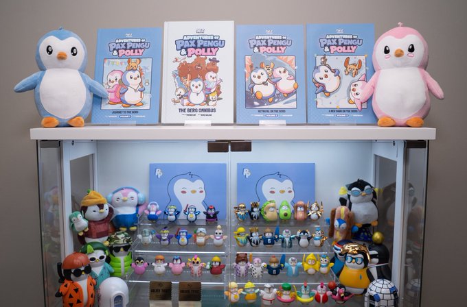A display shelf featuring plush toys and book series titled 'The Adventures of Pax Pengu & Polly', with a glass case below filled with various small penguin figurines.