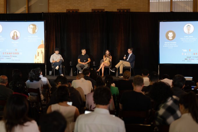 A panel discussion on a stage at an entrepreneurship event called 'STANFORD' with four speakers seated in front of an audience, with large screens displaying their photos and names on either side.