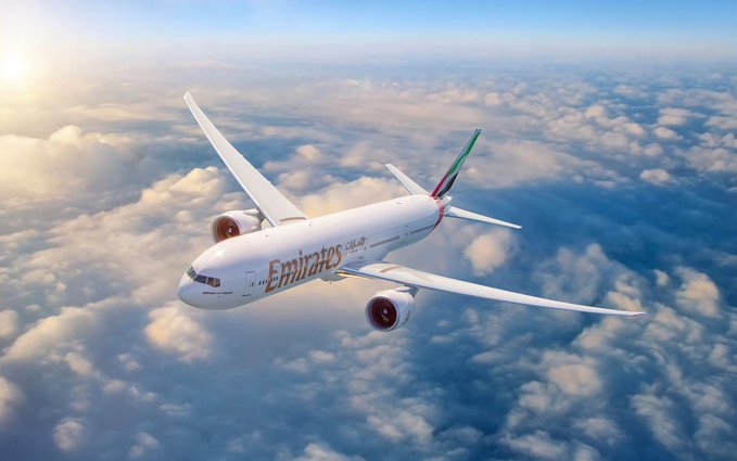 An Emirates passenger jet soars above a layer of clouds in this aerial photograph, with the airline's name and Arabic script clearly visible on the fuselage. The image visually represents Emirates, the UAE's flagship carrier referenced in the breaking news about accepting cryptocurrency for flight bookings.