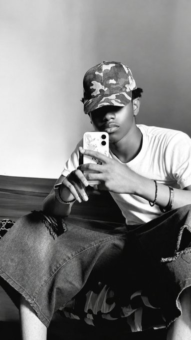 A black-and-white portrait of a young man wearing a camouflage cap and ripped jeans, looking at his smartphone while sitting on a couch.