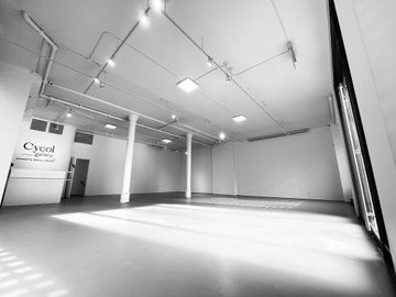 Black and white photograph of an empty warehouse or studio space with white walls, exposed ceiling pipes, and sunlight streaming across the floor; a logo reading 'Cycol Prestige' is visible on the wall.