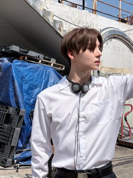A young person with short brown hair poses outdoors wearing a white button-up shirt with headphones around their neck, standing near tarps and metal structures at what appears to be an industrial or filming location. The hashtags #ALPHADRIVEONE, #ALCHA, and related tags in the post suggest this is promotional content for the artist ALD1 and their work.