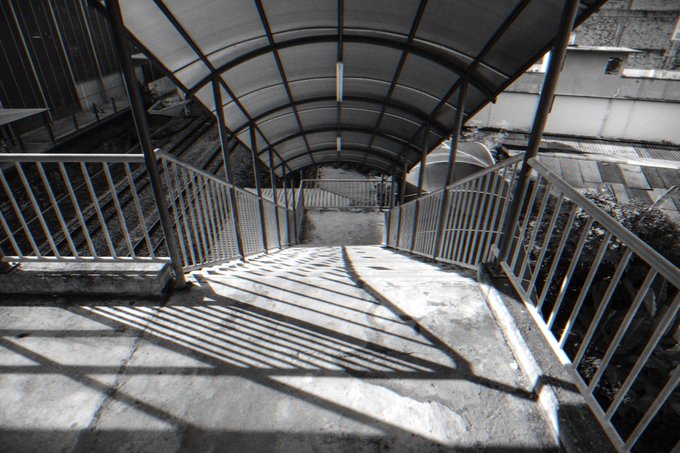 A pedestrian walkway with a curved, arched metal canopy is covered by metal railings on both sides, casting strong geometric shadows on the concrete ground.