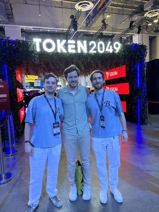 Three men posing together at the TOKEN2049 conference, with TRON branding visible on the entrance display behind them.