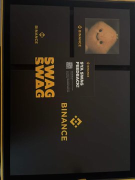 A Binance promotional package labeled 'SWAG BINANCE' containing various branded merchandise including a cap, displayed on a dark background.