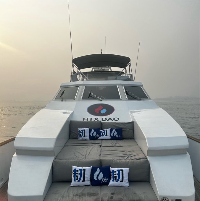 Front view of a white luxury boat named 'HTX DAO' on the water, featuring cushioned seating with blue and white branded pillows at sunset or sunrise.