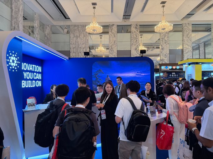 Attendees with backpacks gather at a busy tech conference booth with blue lighting and a sign reading 'IOVATION YOU CAN BUILD ON'.