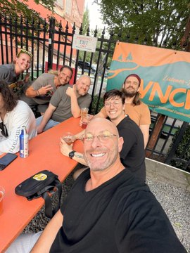 A group of friends gathers around an orange picnic table at an outdoor event, with one man taking a selfie in the foreground while the banner behind them reads "ANCI..." The caption "All right everyone your boy #PROOFOFHUMAN with the free pass from the pregnant wife!!" matches the lively, social atmosphere captured in the image, likely taken at a Bitcoin or cryptocurrency conference as suggested by the mention of #SPX6900.