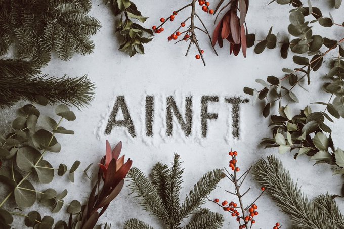 A winter-themed photograph showing the text 'AINFT' written in snow, surrounded by an arrangement of green pine branches, eucalyptus leaves, and red berries in a flat lay composition.