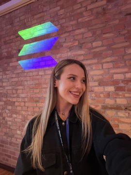 A smiling blonde woman wearing a black jacket and lanyard, posing for a selfie in front of a brick wall featuring a colorful Solana logo.
