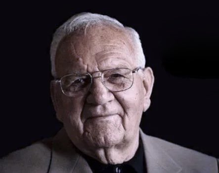 A black and white portrait of an elderly man with white hair and glasses, wearing a suit and tie against a dark background.