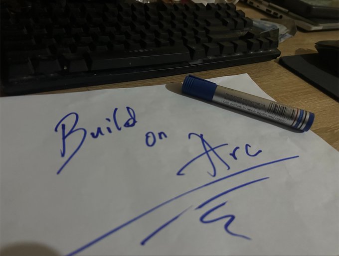 Handwritten text on a piece of paper reads "Build on Arc," with a blue marker resting beside it, aligning with the post's theme of building on @arc.