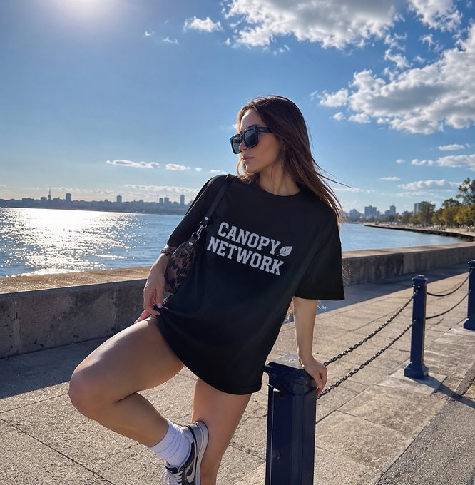 A young woman wearing sunglasses and a black 'CANOPY NETWORK' t-shirt poses by a waterfront railing with a city skyline visible in the distance under a partly cloudy sky.