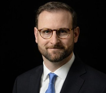 A professional headshot of a bearded man wearing glasses, a dark suit, white shirt, and blue tie, photographed against a black background.