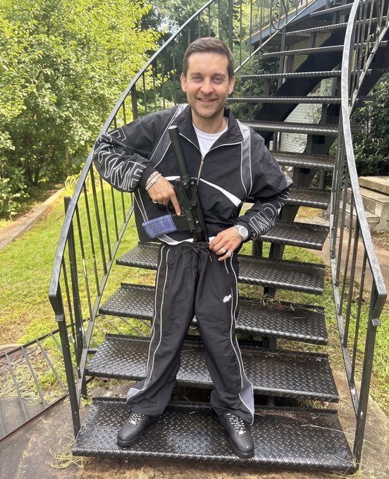 A smiling man in a black Nike tracksuit stands on an outdoor metal spiral staircase while holding what appears to be a rifle or large prop gun.