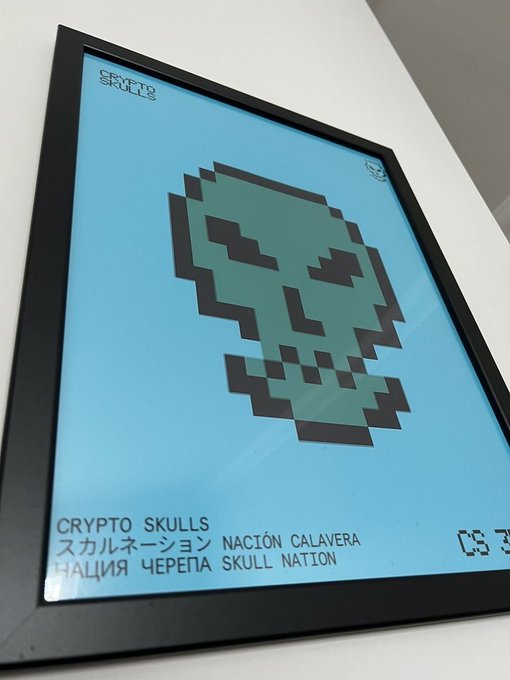 Framed pixel-art print of a teal skull on a light blue background labeled 'CRYPTO SKULLS' with multilingual text 'スカルネーション NACIÓN CALAVERA ЧЕРЕПА SKULL NATION' and edition numbers at the bottom.