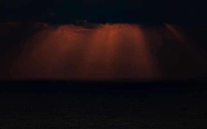 A dramatic sky shows sunbeams (crepuscular rays) breaking through dark clouds at what appears to be sunset or sunrise, casting warm orange light over a dark horizon.