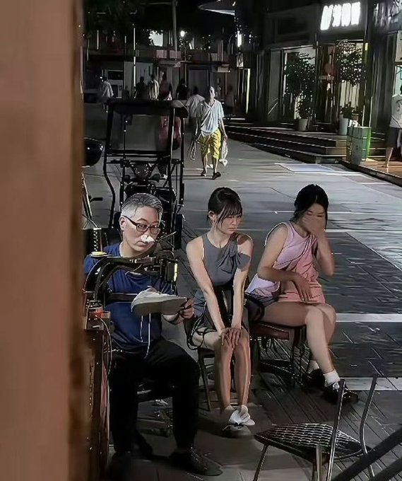 A nighttime street scene showing three people sitting on chairs — a man with glasses reading a book or clipboard, and two women next to him — in what appears to be an outdoor café or pedestrian street setting.