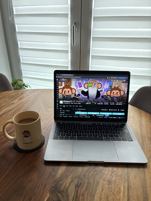 A laptop sits open on a wooden table displaying the 'Chimpers NFT' collection, accompanied by a beige mug with a cartoon character design and a small potted plant near a window with white blinds.