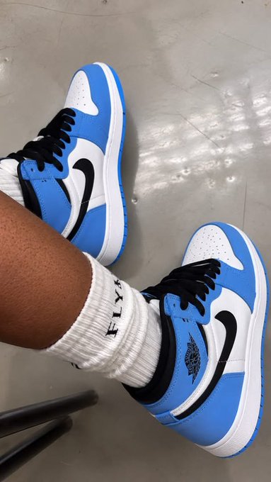 A pair of feet wearing blue, white, and black Air Jordan 1 sneakers with white crew socks on a tiled floor.