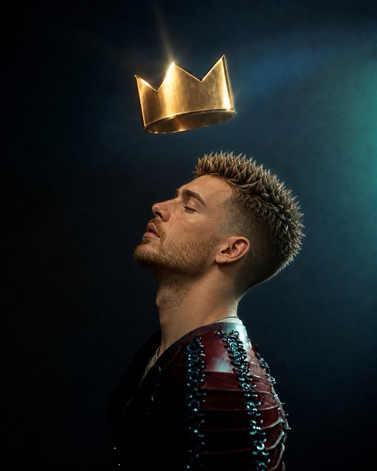 A man in dark, scaled armor stands with his eyes closed and head tilted upward as a golden crown floats above him.